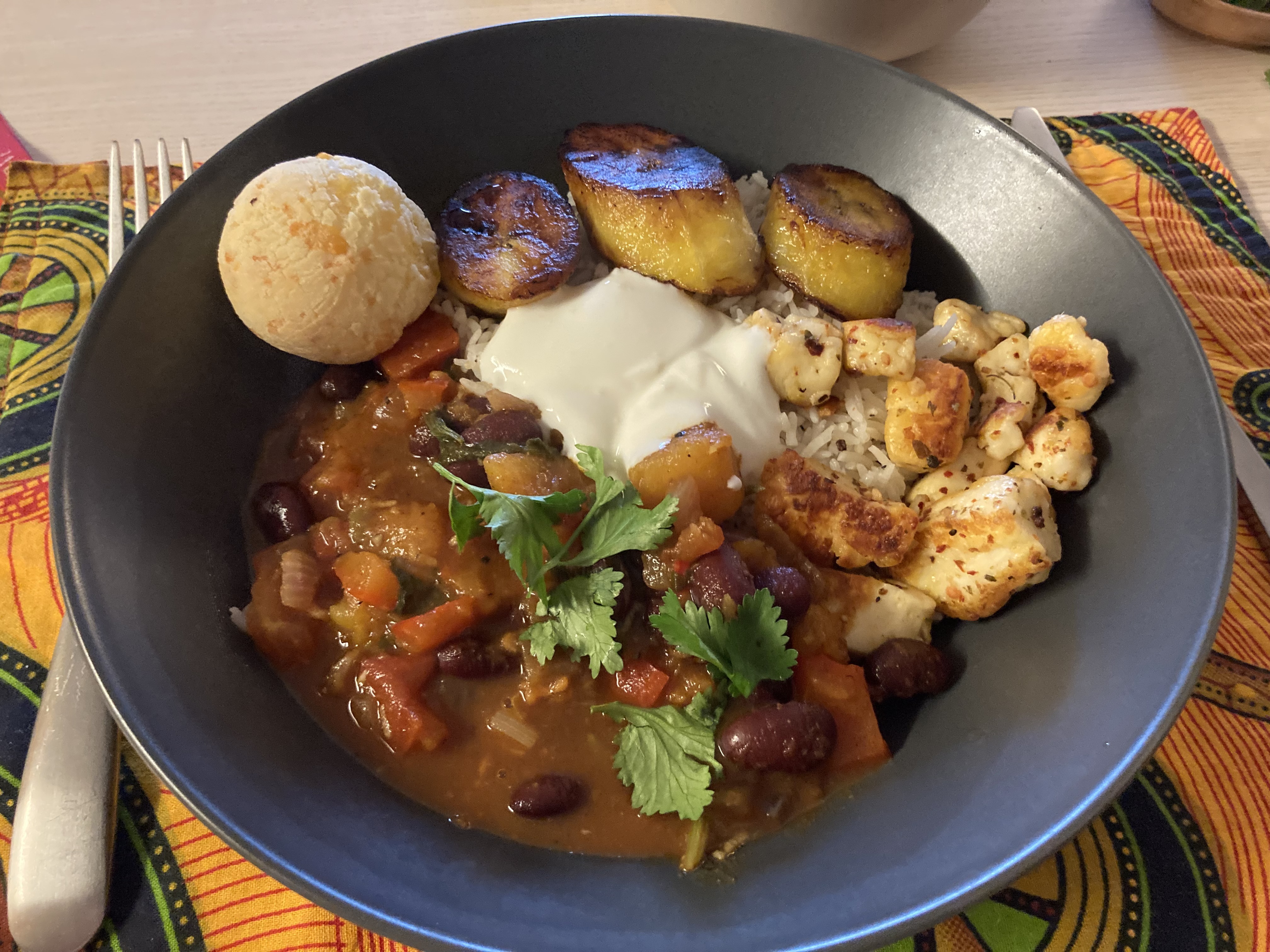 A luscious looking deep red and brown vegetable bean stew with halloumi, fried plantains, bright green coriander, and pao de queijo cheese puff ball.